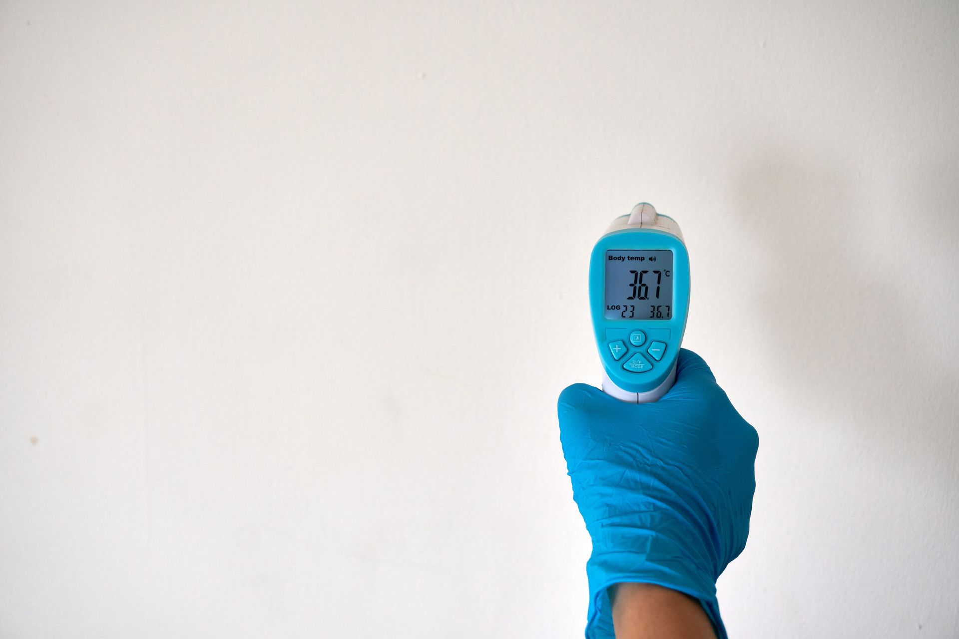 A hand in blue gloves holding an infrared thermometer against a plain background, signifying safety and health measures.